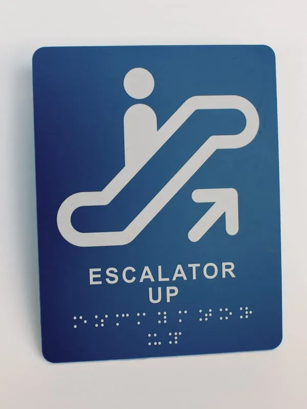 A blue ADA escalator-up sign with pictogram, lettering, and braille